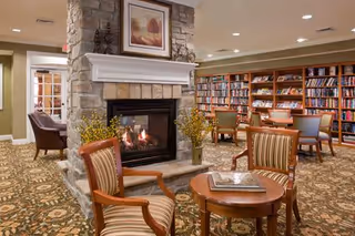 Cozy senior living facility common area with a stone fireplace in the center, surrounded by comfortable chairs and small tables. Bookshelves filled with books line the back wall, and the room is warmly lit with recessed ceiling lights.