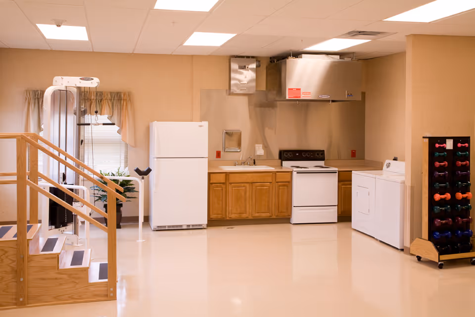 A room featuring a small kitchen area with a white refrigerator, stove, and washing machine. There are wooden cabinets below the countertop and a stainless steel range hood above the stove. To the left, there is a wooden staircase with handrails and exercise equipment near a window with beige curtains. On the right side, there is a rack holding colorful dumbbells.