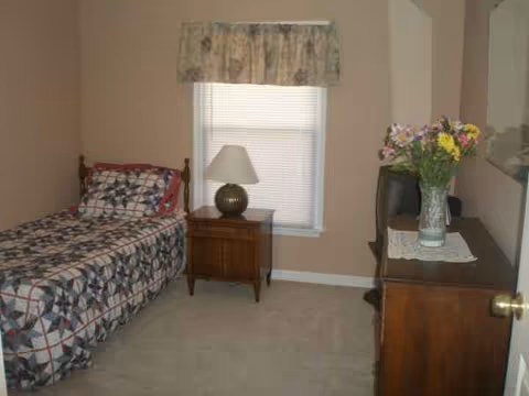 A small senior living bedroom with a single bed, nightstand, dresser, TV, and flowers.