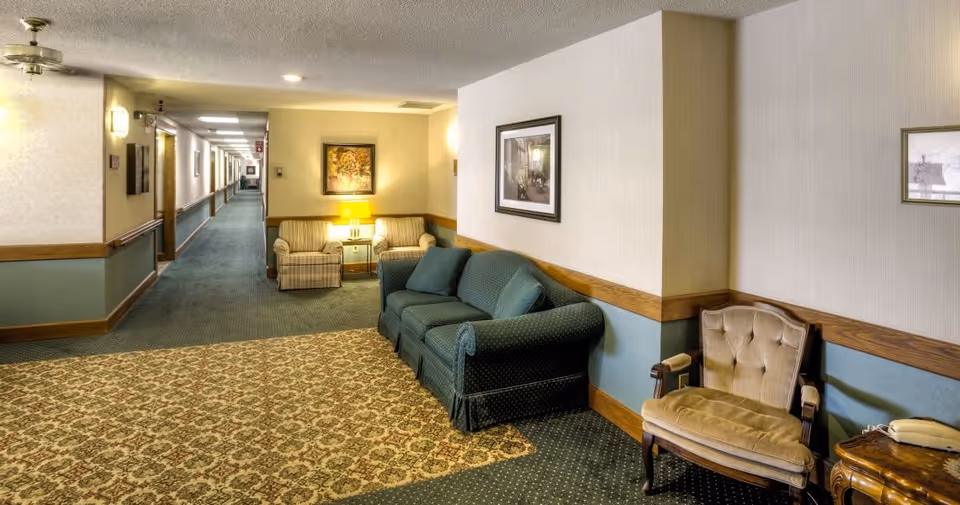 A hallway in a senior living facility with patterned carpet and walls painted in beige and blue with wooden trim. The hallway features a green upholstered sofa, a beige armchair, two striped armchairs, a side table with a lamp, framed artwork on the walls, and ceiling lights. The hallway extends into the distance with multiple doors along the left side.