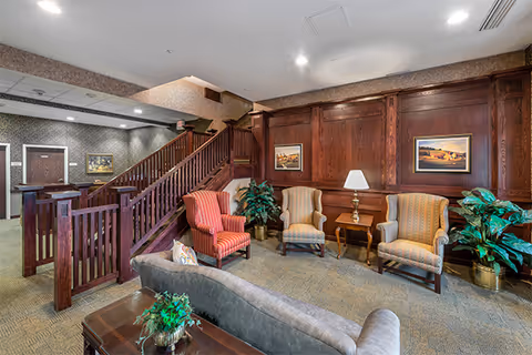 Seating area in a senior living lounge with armchairs, a sofa, side tables and lamp, potted plants, framed art, and a wooden staircase.