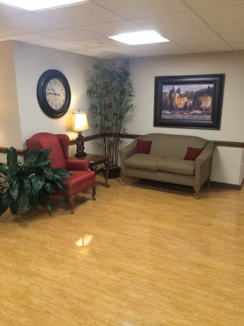 A cozy sitting area with a red upholstered armchair, a small wooden side table with a lamp, a green sofa with two red pillows, a large wall clock, a framed painting, and two potted plants. The floor is wooden and the walls are painted beige.