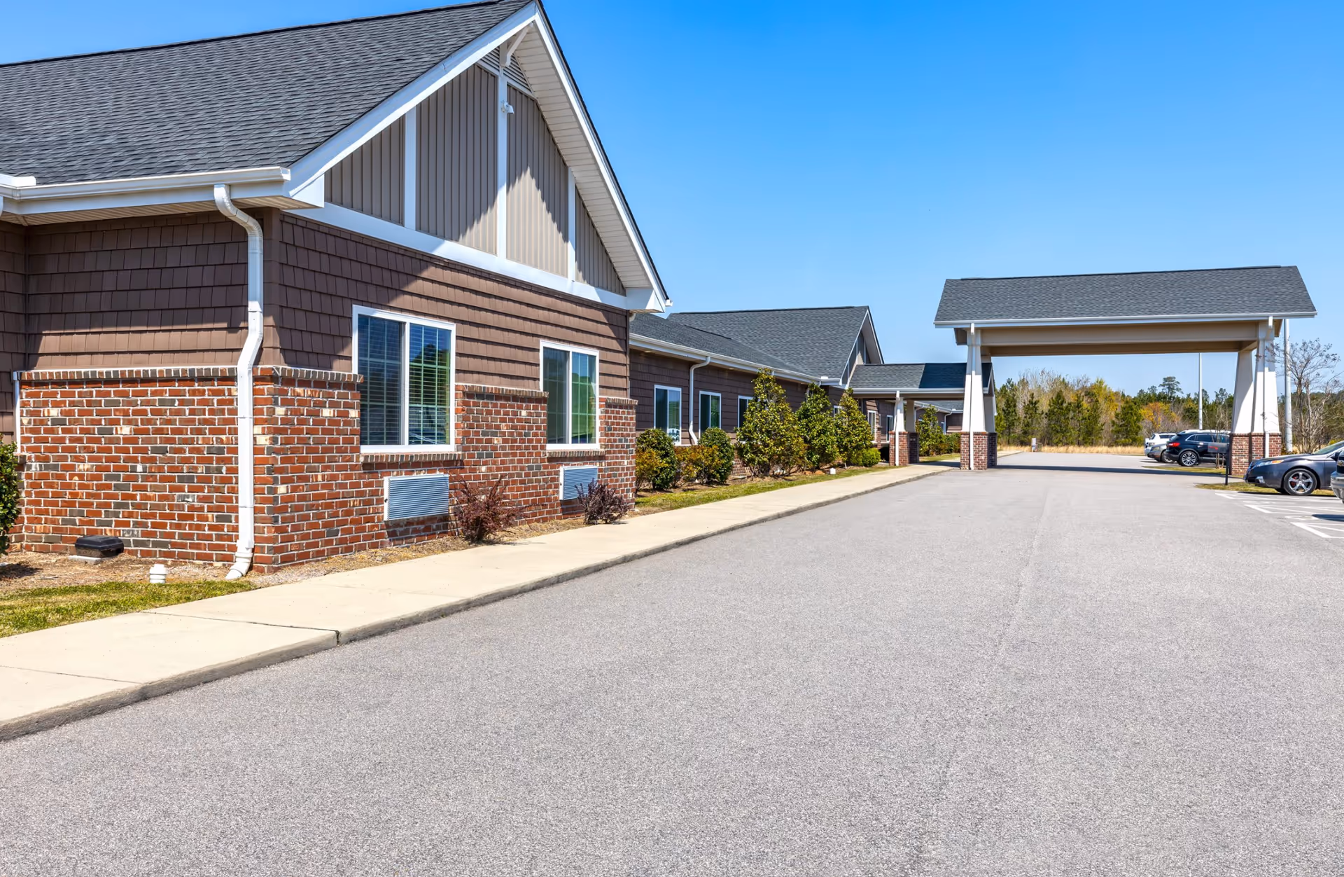 Exterior view of a senior living facility named Gates House showing a long building with brown siding and brick accents, multiple windows, a covered entrance with a pitched roof, a paved driveway, parked cars, and clear blue sky.