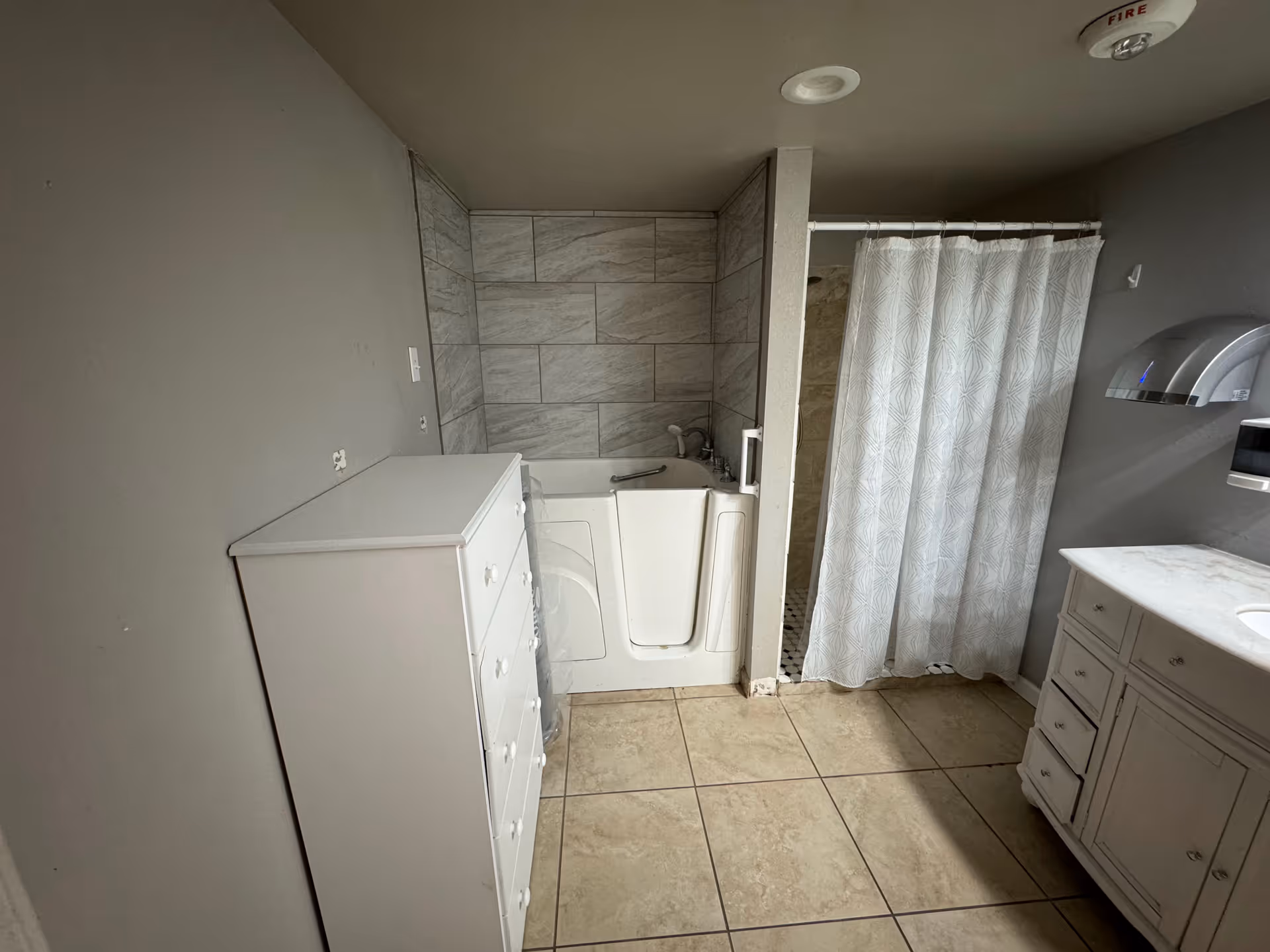 A bathroom with beige tiled floor and gray walls. There is a white walk-in bathtub with a tiled wall surround, a white shower curtain on a rod, a white chest of drawers, and a white vanity with a sink. A hand dryer is mounted on the wall above the vanity.