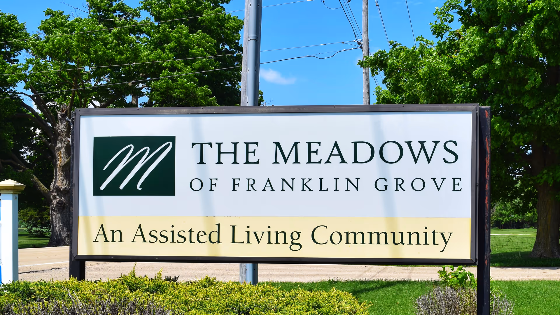Outdoor sign for The Meadows of Franklin Grove, an assisted living community, with green trees and blue sky in the background.