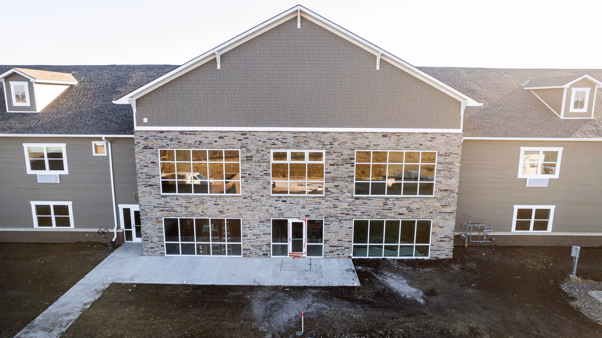 Front exterior view of a two-story building with a stone facade on the lower half and gray siding on the upper half. The building has multiple large windows and a central glass door. The surrounding ground appears to be under construction or landscaping.