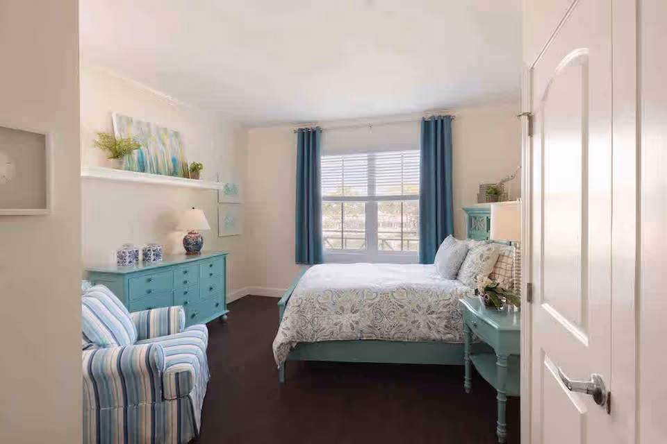 A bright and cozy bedroom with a large window covered by blue curtains. The room features a bed with patterned bedding, a matching blue nightstand with a plant, a blue dresser with decorative items and a lamp, and a striped armchair. The walls are light-colored, and there is a shelf with artwork and plants above the dresser.