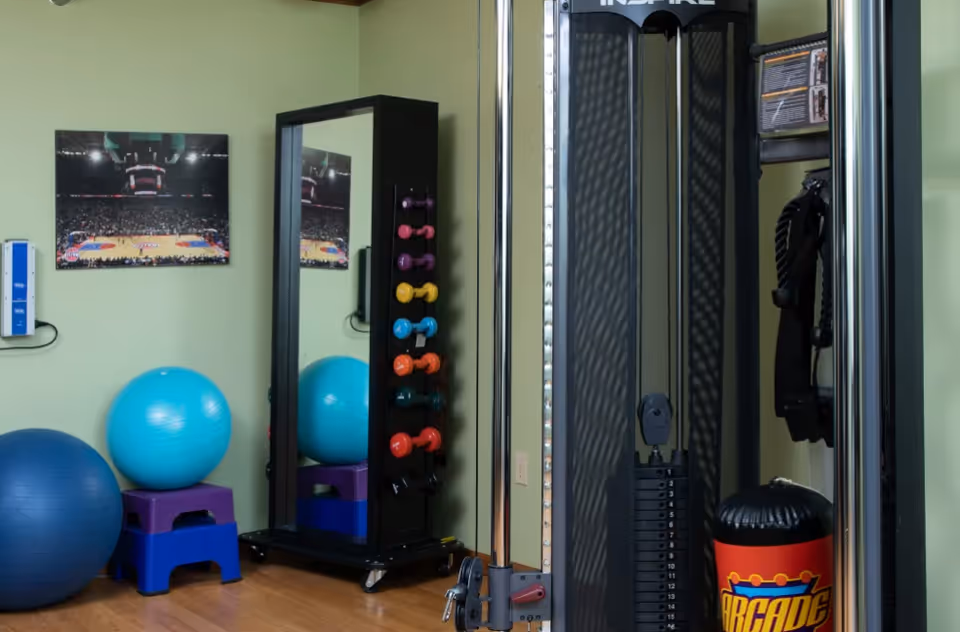 A fitness room with exercise equipment including a weight machine, colorful dumbbells on a rack, two large exercise balls, a mirror, and a framed picture of a basketball court on the wall.