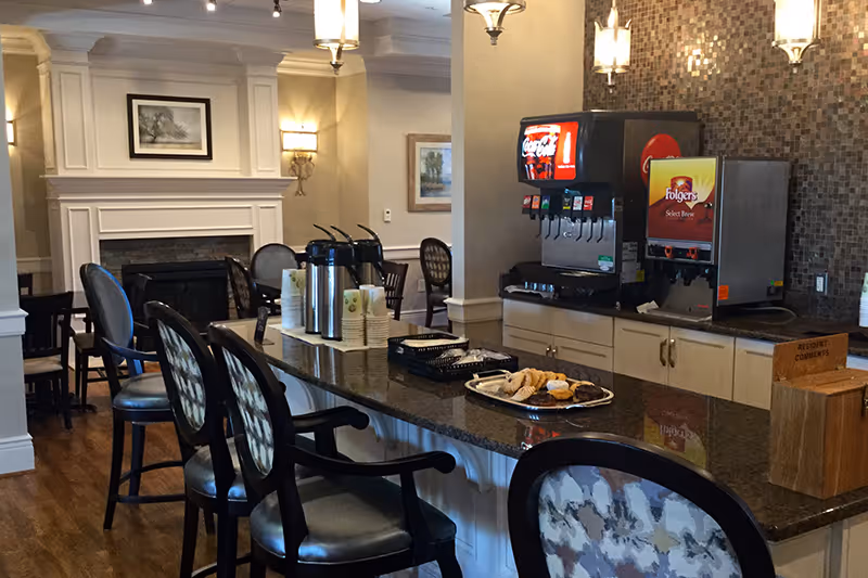 Interior view of a senior living facility common area with a granite countertop island featuring coffee dispensers, a soda fountain, and a tray of pastries. Several cushioned chairs with patterned backs are arranged around the island. In the background, there is a fireplace with a framed picture above it and additional seating.