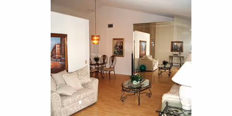 A cozy living room area with beige patterned sofas, a glass coffee table with decorative items, a side table with a lamp, and a small dining area with a round table and chairs in the background. The room has wooden flooring and framed artwork on the walls.