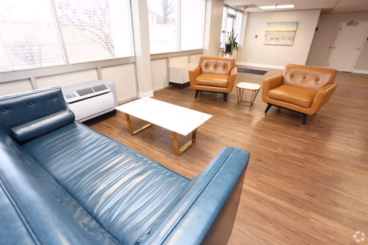 A bright and spacious common area with large windows along one wall. The room features a blue leather sofa, two tan leather armchairs, a white marble coffee table with gold legs, and a small side table. The floor is wood, and there is a painting on the far wall near a doorway.