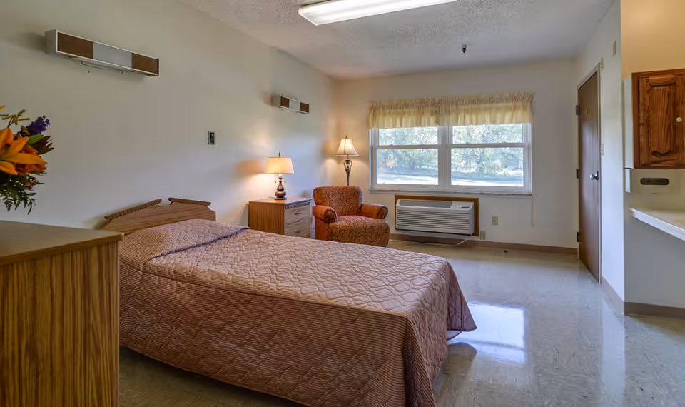 A simple senior living bedroom with a single bed, nightstand and lamp, upholstered armchair by a window and a wall air conditioner.