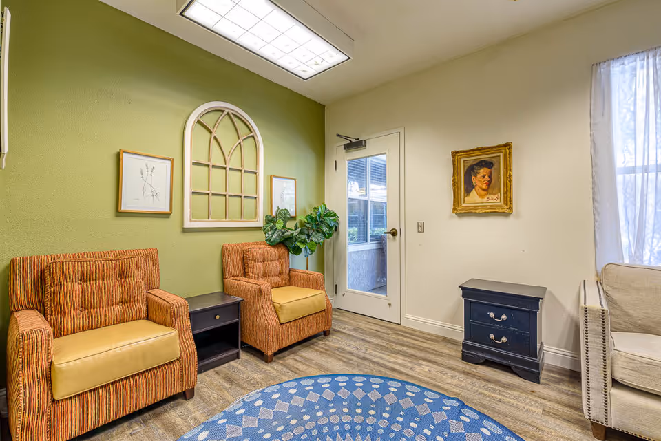A cozy sitting area in a senior living facility with two striped orange armchairs with yellow cushions, a small dark wooden side table between them, a green accent wall with framed artwork and a decorative window frame, a door with a window leading outside, a blue patterned round rug on the wooden floor, a small dark blue chest of drawers, a beige armchair, and a framed portrait on the wall.
