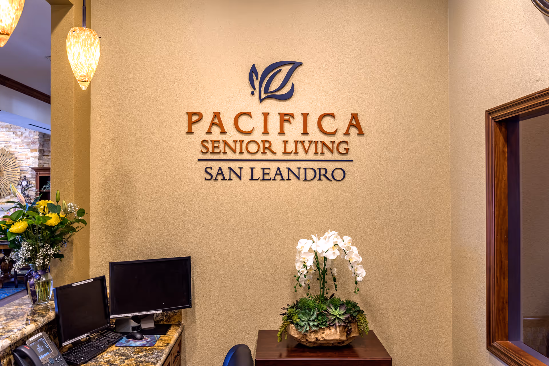 Reception area of Pacifica Senior Living San Leandro with a wall sign displaying the facility name, two computer monitors on a granite countertop, a telephone, a vase with yellow flowers, and a decorative arrangement of white orchids and green succulents on a wooden table.