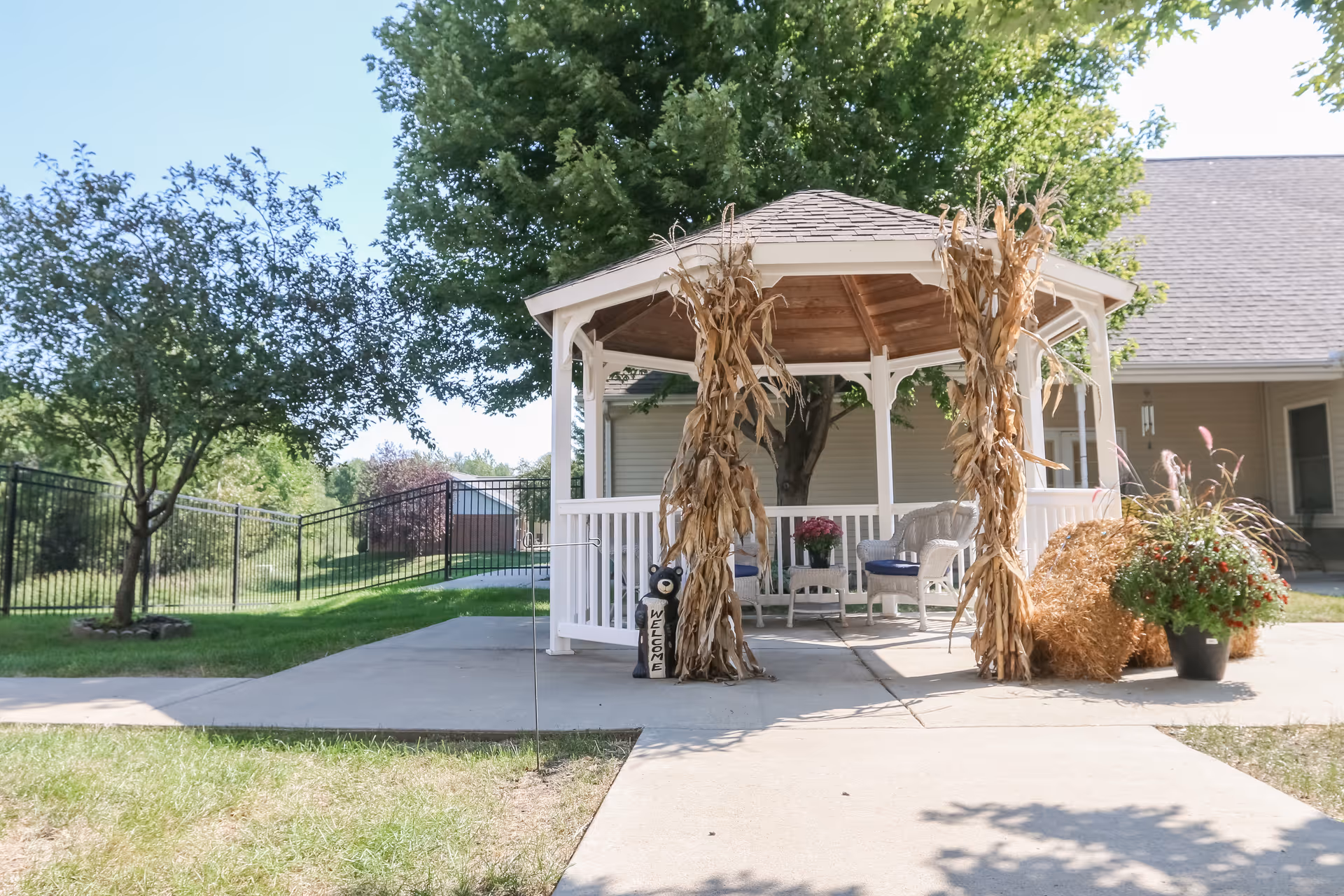 A white wooden gazebo decorated with dried corn stalks and a small welcome sign, situated on a concrete patio surrounded by green grass and trees. There are white wicker chairs and a small table with a flower pot inside the gazebo. A building with beige siding and a gray roof is visible in the background.