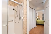 View down a small hallway with a shower stall on the left, closet doors, and a bedroom with a green bed in the background.