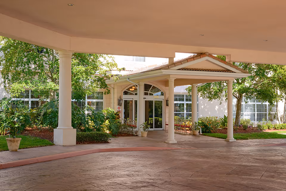 Covered porte-cochere entrance with columns, potted plants, and landscaping at the front of a residential building.