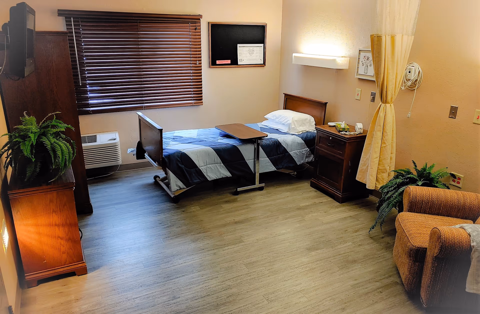 A neatly arranged bedroom in a senior living facility with a single bed covered in a blue and gray checkered blanket, a wooden nightstand with a lamp and some items on it, a wooden cabinet with a green plant on top, a brown armchair with a blanket draped over the arm, a window with closed wooden blinds, and a privacy curtain partially drawn to the side.