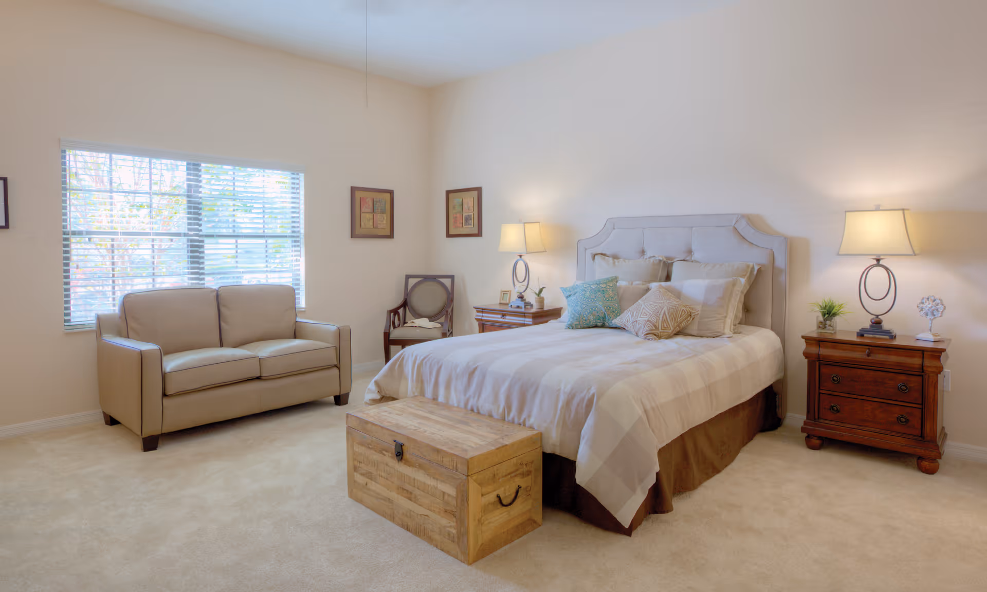 Bright furnished bedroom with a bed, wooden nightstands and lamps, a trunk at the foot of the bed, and a loveseat by the window.