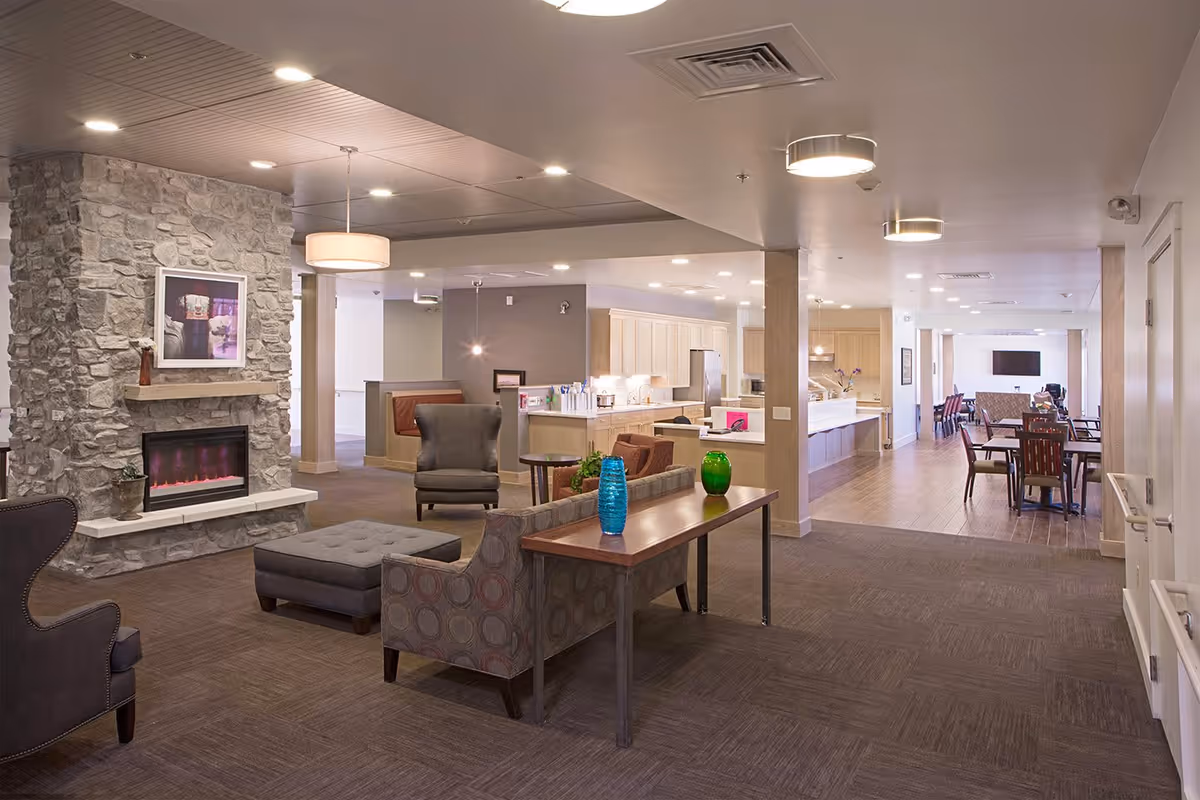 Interior view of a senior living community common area featuring a stone fireplace with a framed picture above it, comfortable seating including armchairs and a sofa, a wooden table with decorative vases, and a kitchen and dining area in the background with tables and chairs arranged neatly.