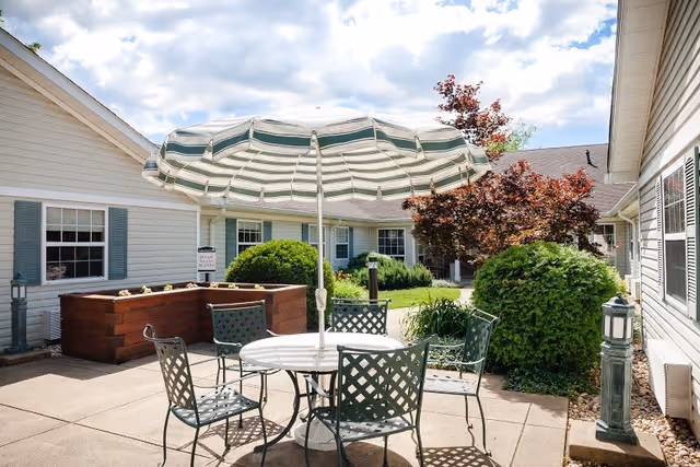 Outdoor patio area with a round white table and four green metal chairs under a large green and white striped umbrella. Surrounding the patio are bushes, a small tree with red leaves, and beige buildings with windows and blue shutters. The sky is partly cloudy.