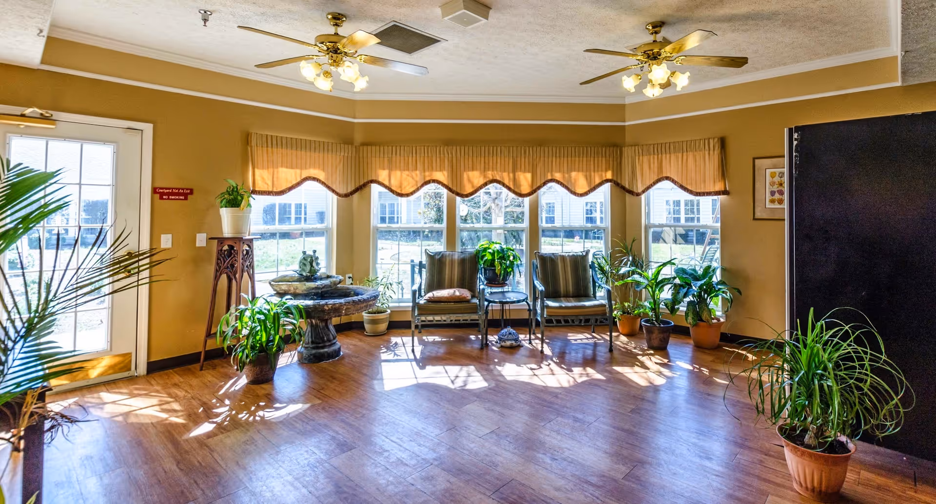 Sunlit seating area with chairs, potted plants, a decorative table, and ceiling fans in a senior living facility lounge.