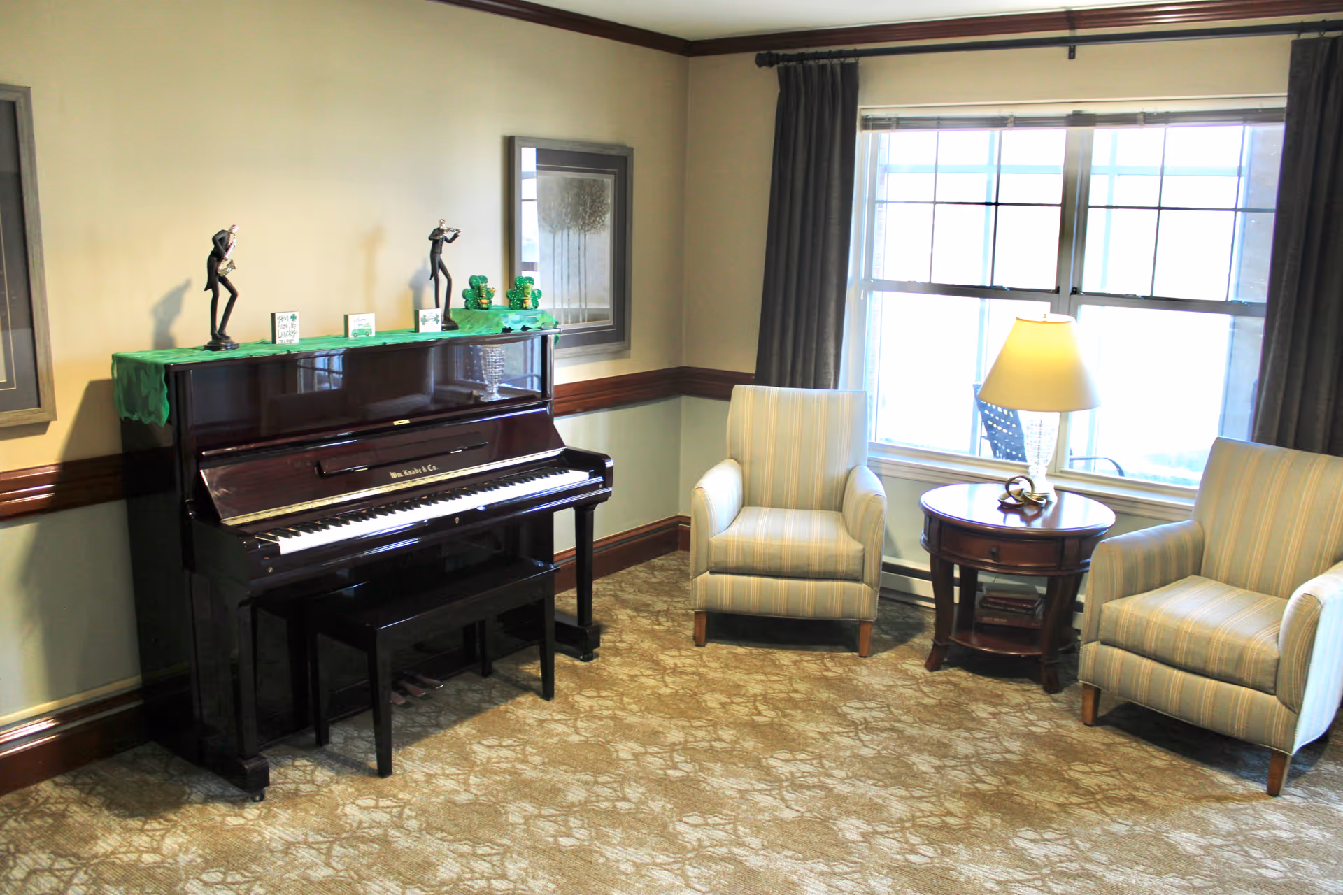 A cozy interior room featuring a dark upright piano with decorative figurines and cards on top, two striped armchairs, a round wooden side table with a lamp, and a large window with curtains letting in natural light.