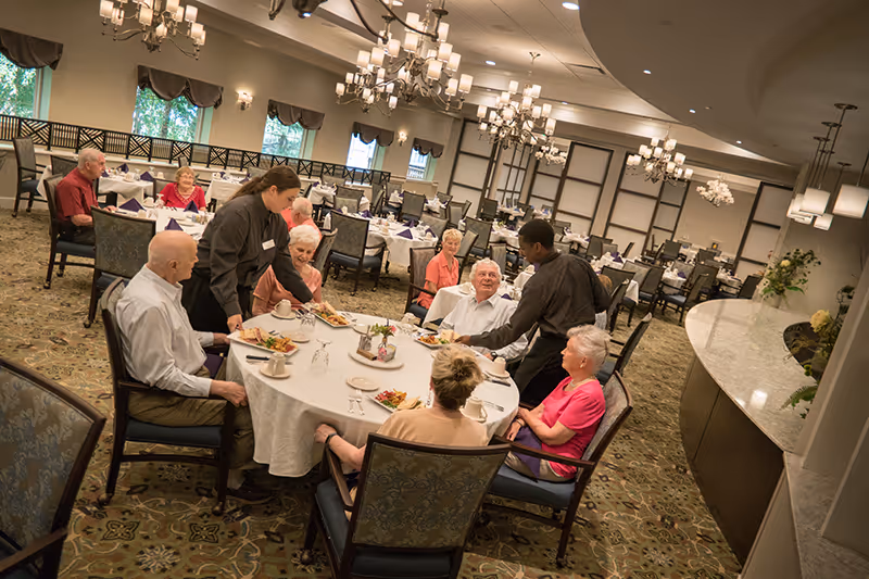 A group of elderly people seated around a large round dining table in a spacious, well-lit dining room. Two staff members are serving food and drinks to the residents. The room has multiple tables with white tablecloths, chairs, chandeliers, and large windows with curtains.