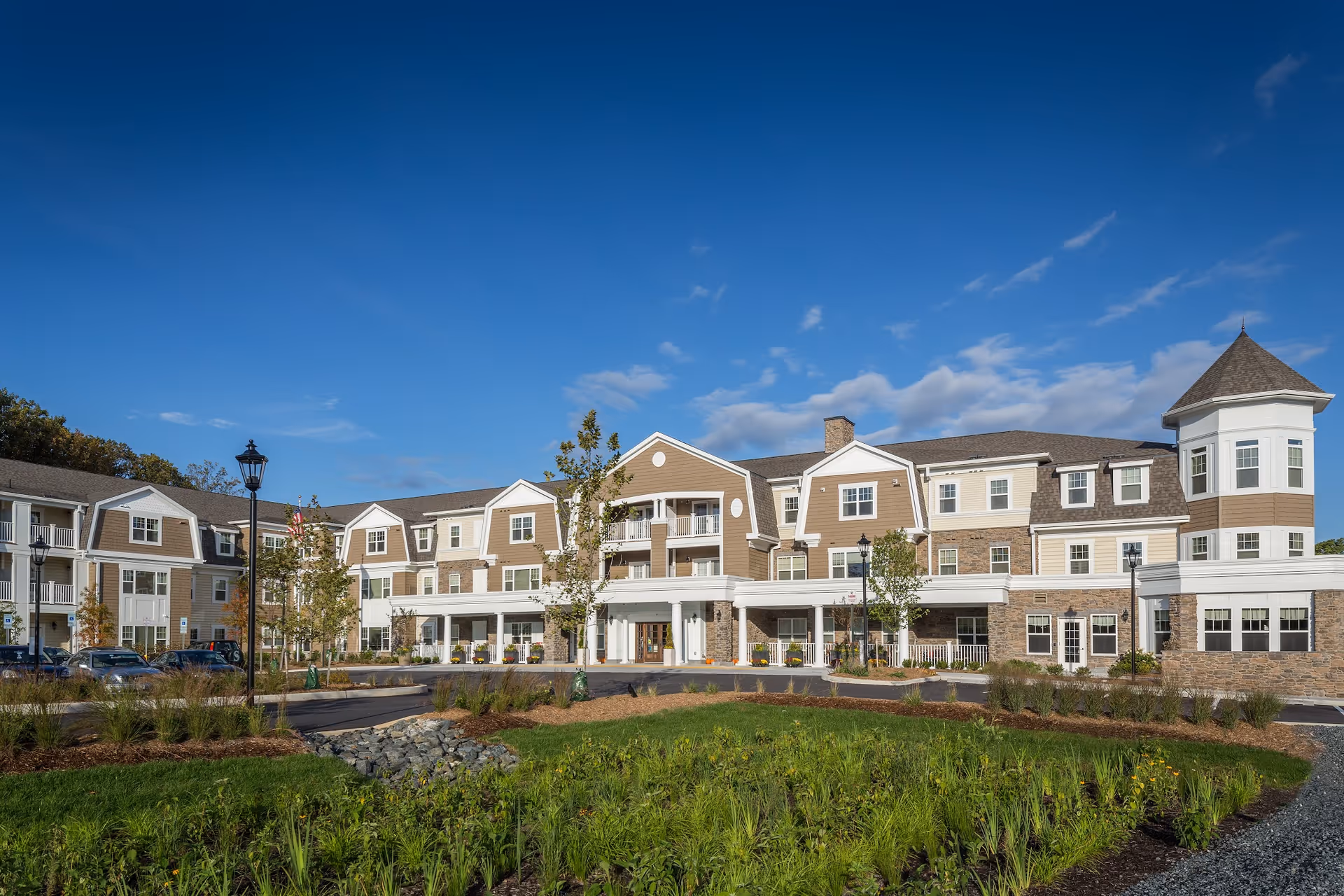 Exterior view of Brightview Rolling Hills senior living facility showing a large, multi-story building with beige and white siding, stone accents, multiple windows, and a turret on the right side. The building is surrounded by landscaped greenery and a clear blue sky.