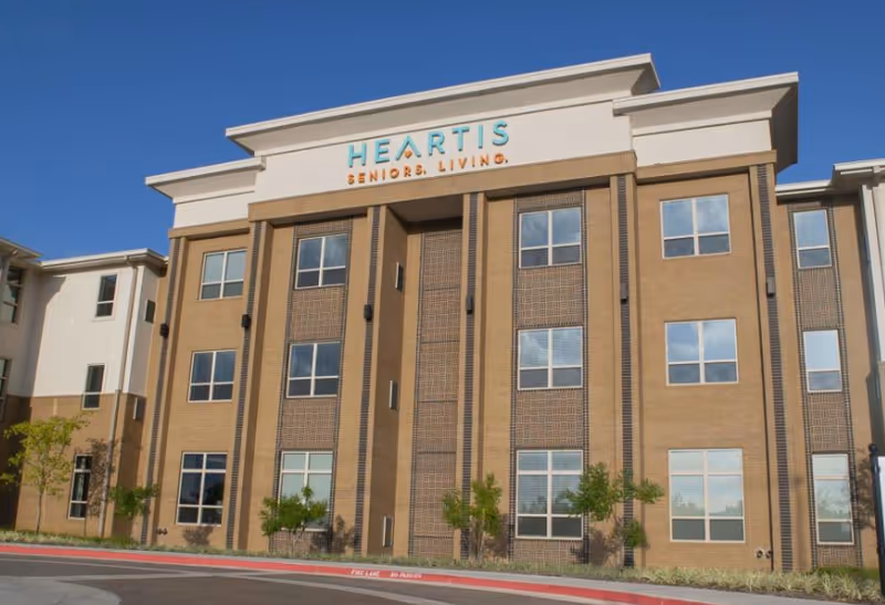 Exterior view of a multi-story senior living facility building with large windows and the sign 'HEARTIS SENIORS LIVING' at the top center. The building is surrounded by small trees and landscaping under a clear blue sky.