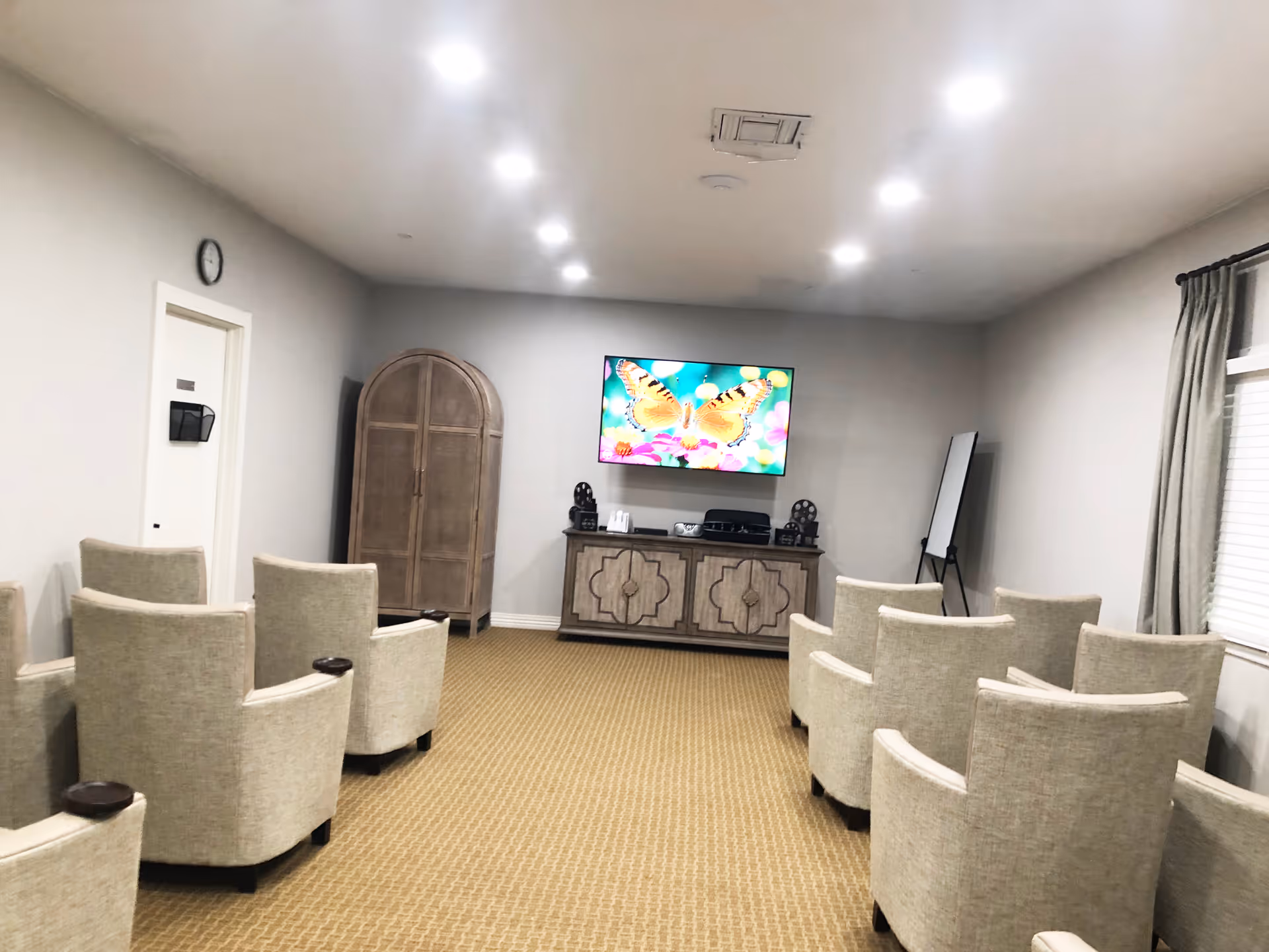 A small meeting or media room with two rows of beige upholstered chairs facing a wall-mounted flat screen TV displaying a butterfly image. Below the TV is a wooden cabinet with decorative patterns, and to the left is a tall wooden armoire. There is a whiteboard on an easel in the corner near a window with closed blinds and curtains. The room has beige carpet and white walls with recessed ceiling lights.