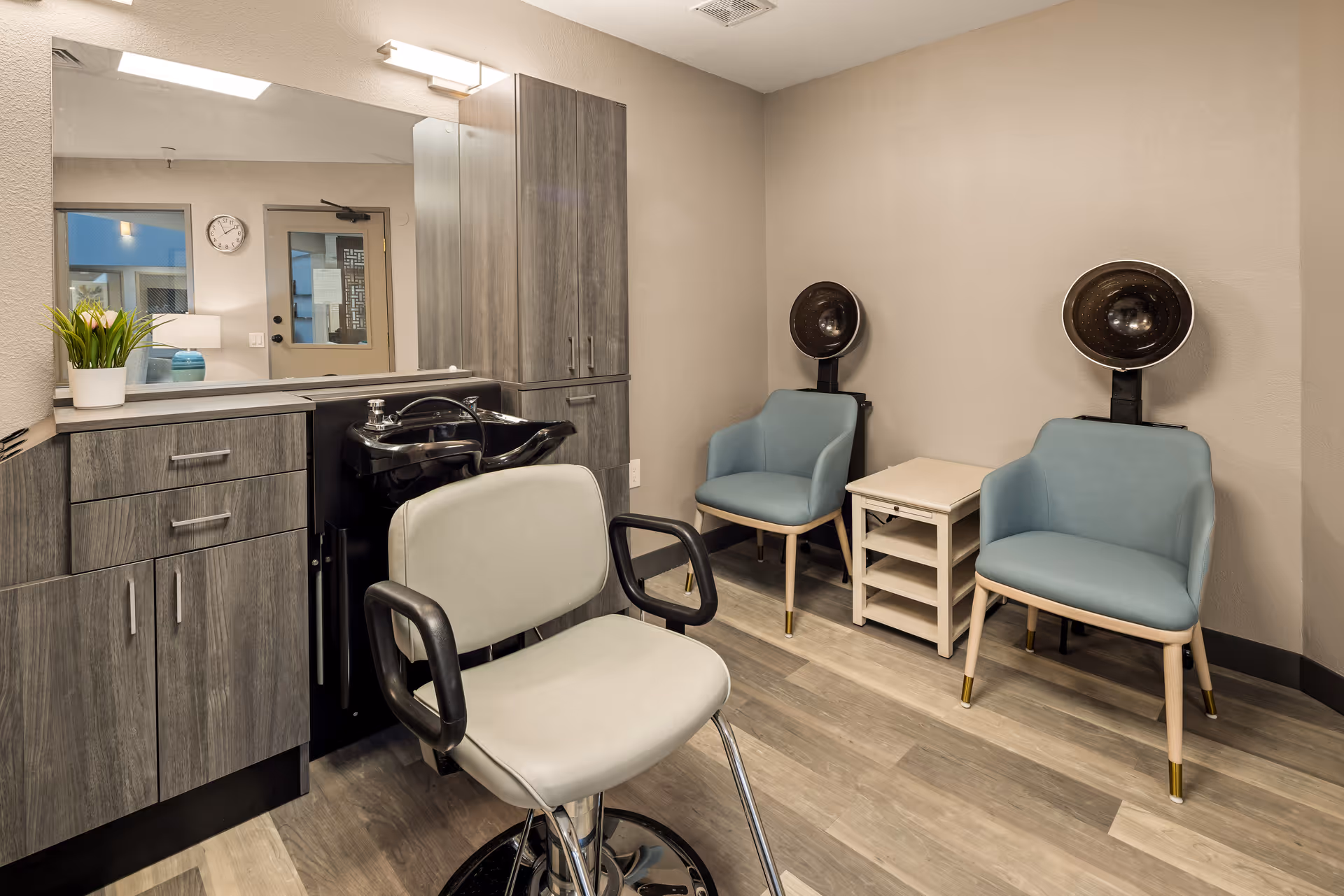 A small salon area in a senior living facility featuring a styling chair in front of a black hair washing sink, gray wooden cabinets with a potted plant on the counter, and two blue chairs with hair drying hoods next to a small white side table. A large mirror is mounted on the wall above the cabinets.