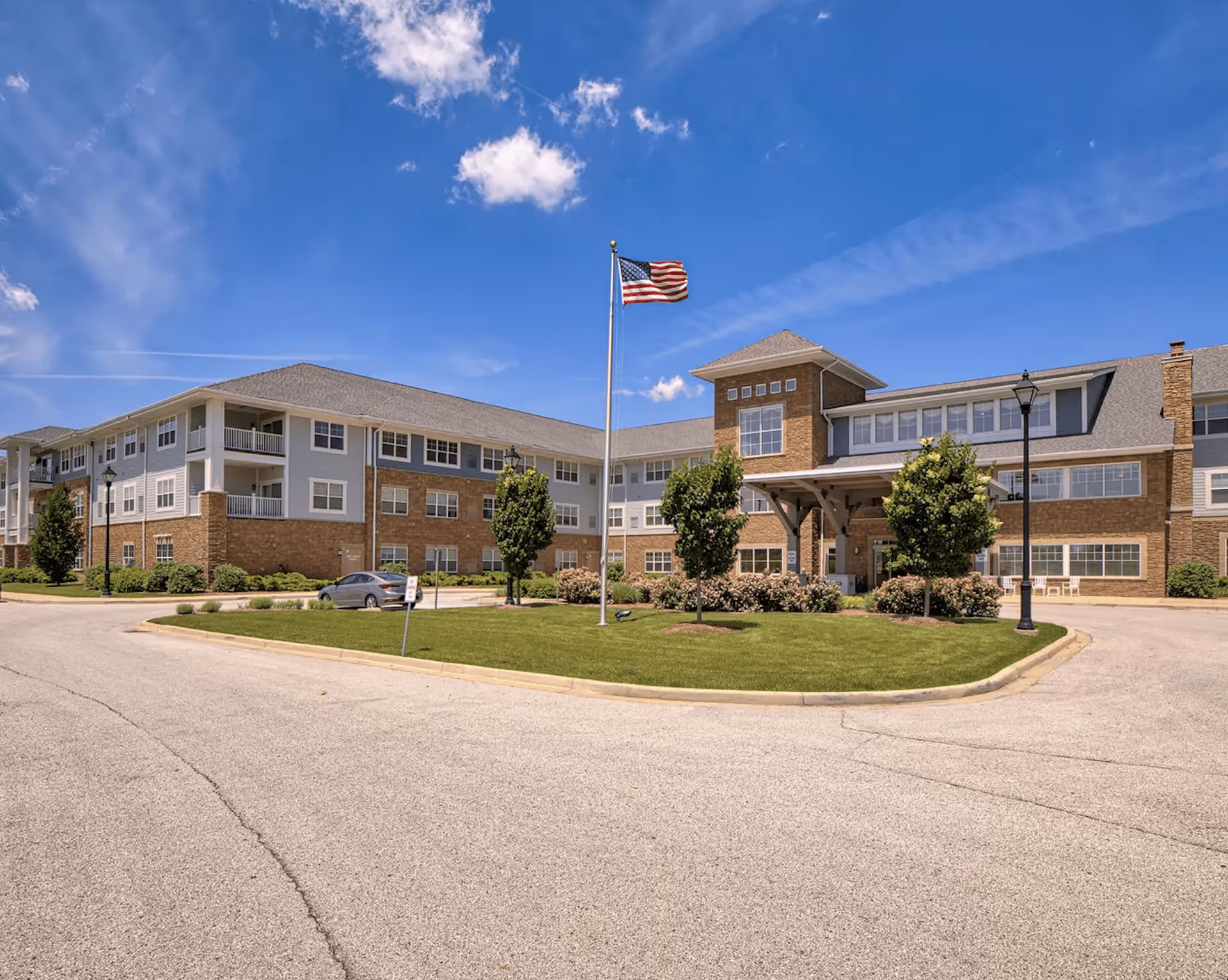 Exterior view of a large senior living facility building with multiple windows and balconies under a blue sky with some clouds. There is a circular driveway with a grassy island in the center featuring an American flag on a flagpole and several small trees and bushes.