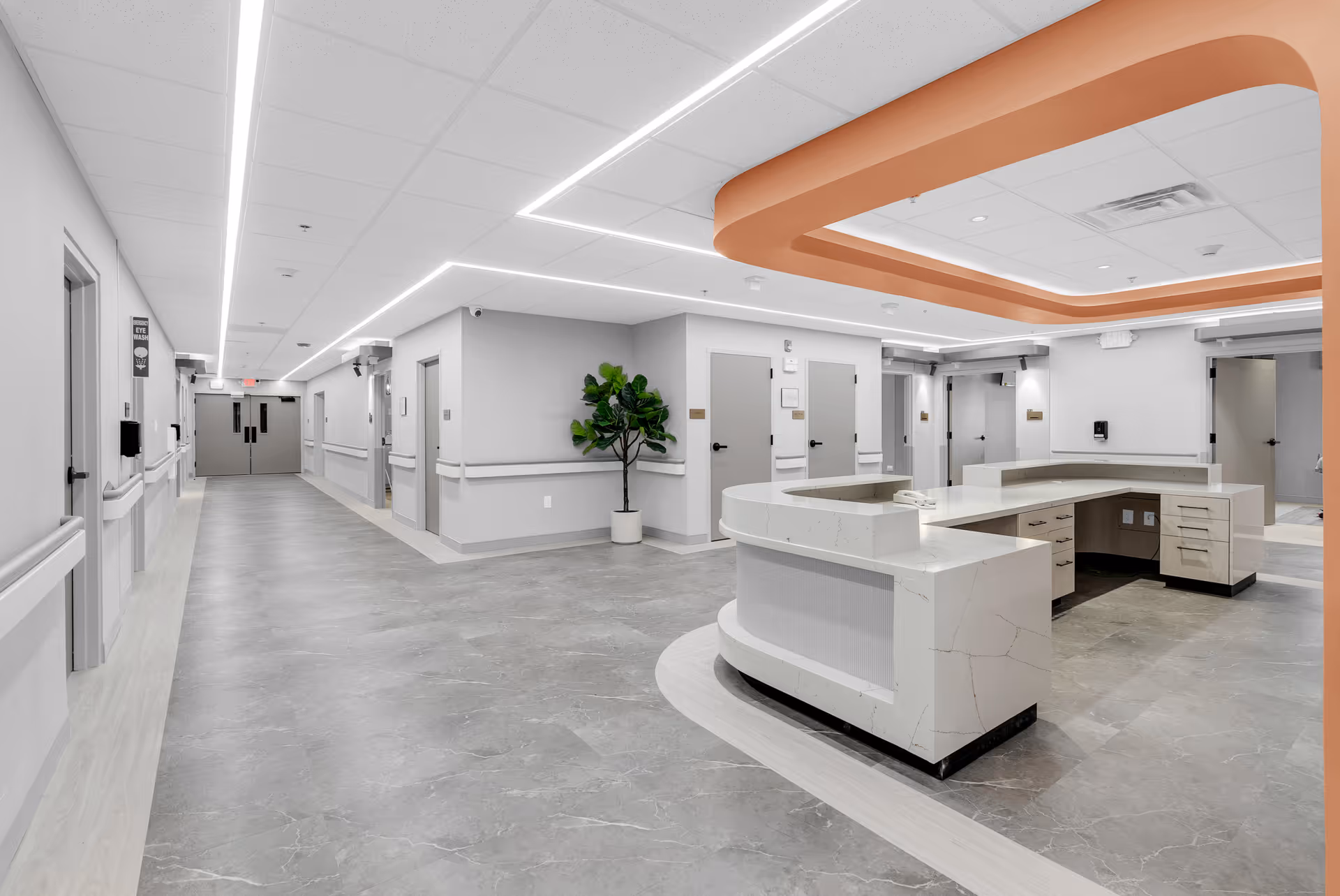 A modern nursing facility hallway with a large, curved reception desk on the right side. The floor is covered with gray tiles, and the walls are painted light gray with white trim. There are multiple closed doors along the hallway and a potted plant near the corner. The ceiling features recessed lighting and a distinctive orange accent around the reception area.