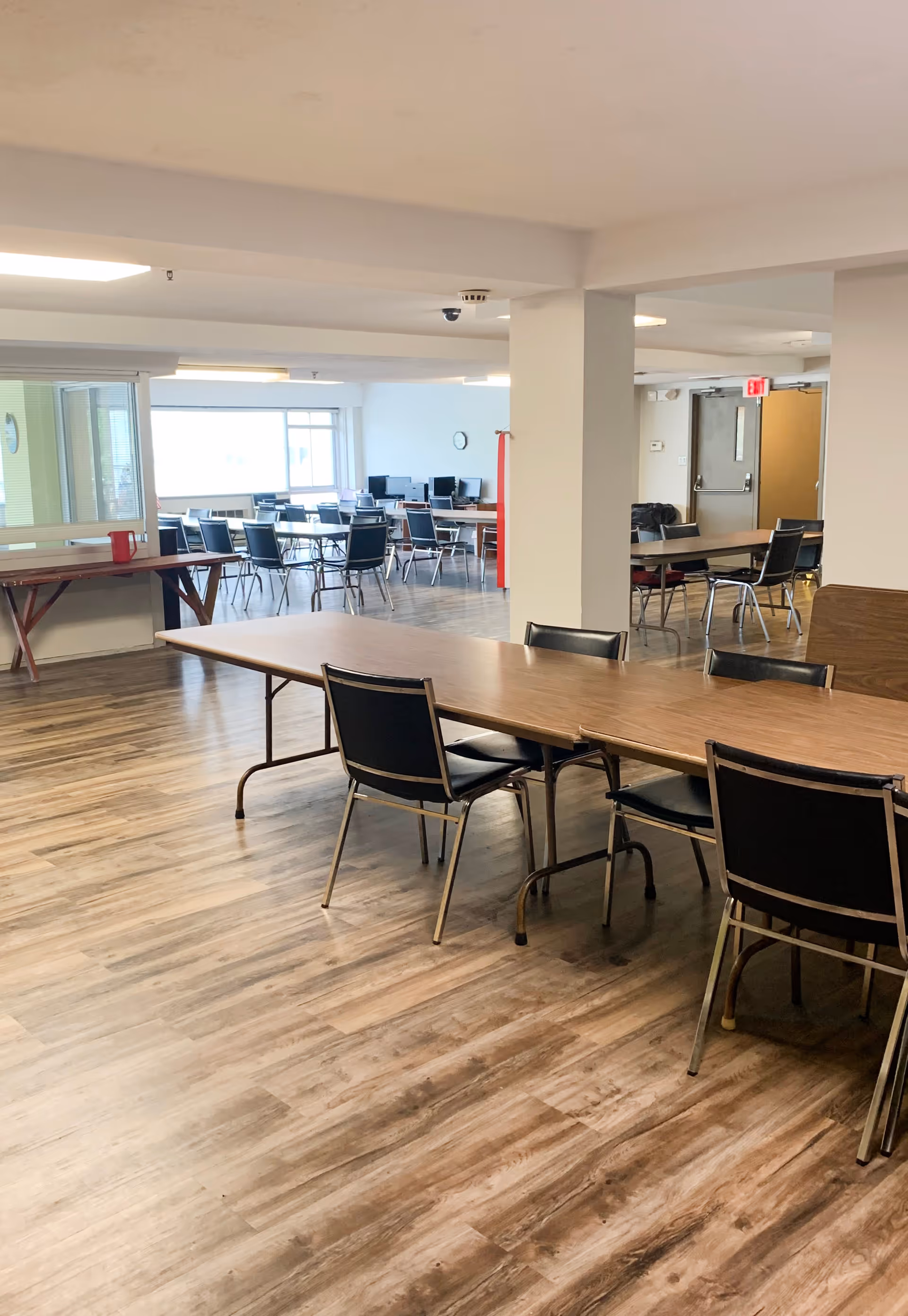 Large communal dining/activity room with long folding tables and chairs on wood-look flooring and computer stations visible in the background.