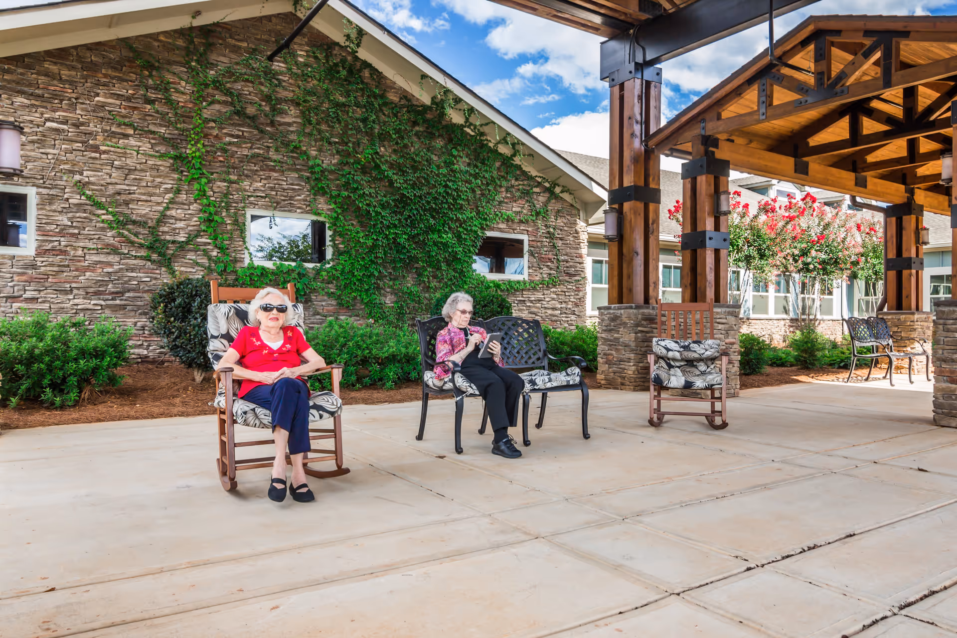 Two elderly women sitting outdoors on cushioned chairs under a wooden pergola structure. One woman is seated in a rocking chair wearing sunglasses and a red shirt, while the other is sitting on a metal bench looking at a tablet. The background shows a stone building wall with green ivy and bushes, and a partly cloudy blue sky.