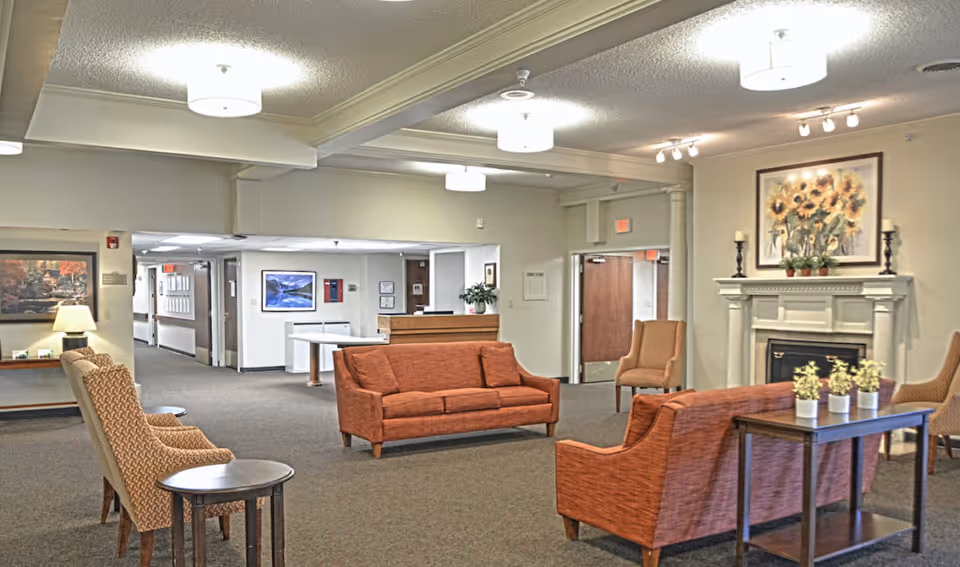 A spacious senior living facility common area with orange upholstered sofas and chairs arranged around a dark wooden table with small potted plants. There is a white fireplace with a painting of sunflowers above it, and soft ceiling lights illuminate the room. The walls are light-colored, and there are hallways and doors visible in the background.