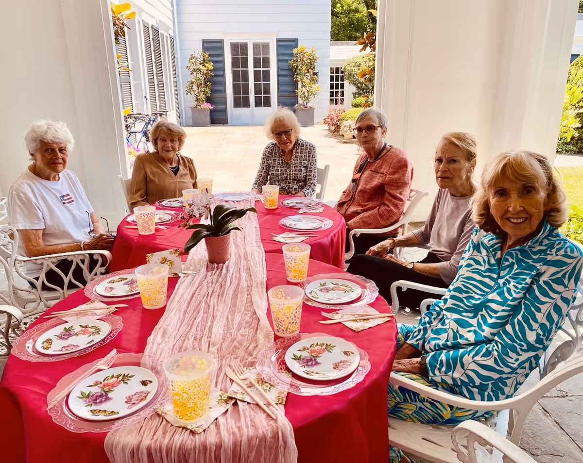 Six elderly women sitting around a table set for a meal with floral plates, yellow patterned cups, and a pink table runner, on a covered patio with white pillars and a view of a garden and building exterior in the background.