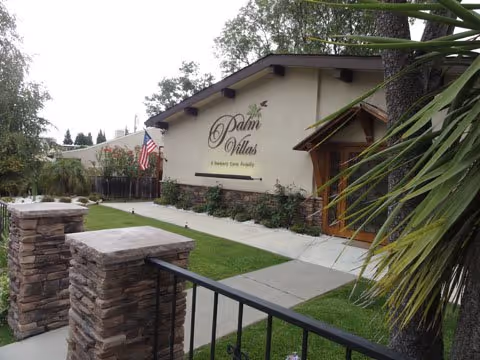 Exterior view of Palm Villas Memory Care facility showing a beige building with a wooden entrance door, a sign reading 'Palm Villas A Memory Care Facility', a walkway, green lawn, stone pillars, and an American flag.