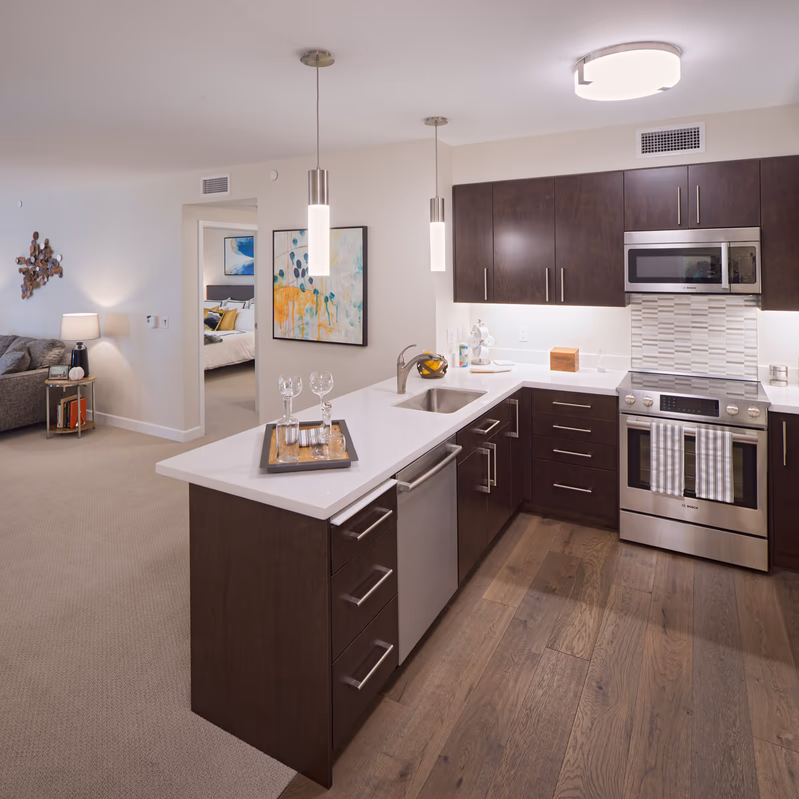 Modern kitchen with dark wood cabinets, white countertops, stainless steel appliances including a stove and microwave, and a sink on the island. Two pendant lights hang above the island, which has a tray with glassware. In the background, a living area with a couch and a bedroom with a bed are visible.