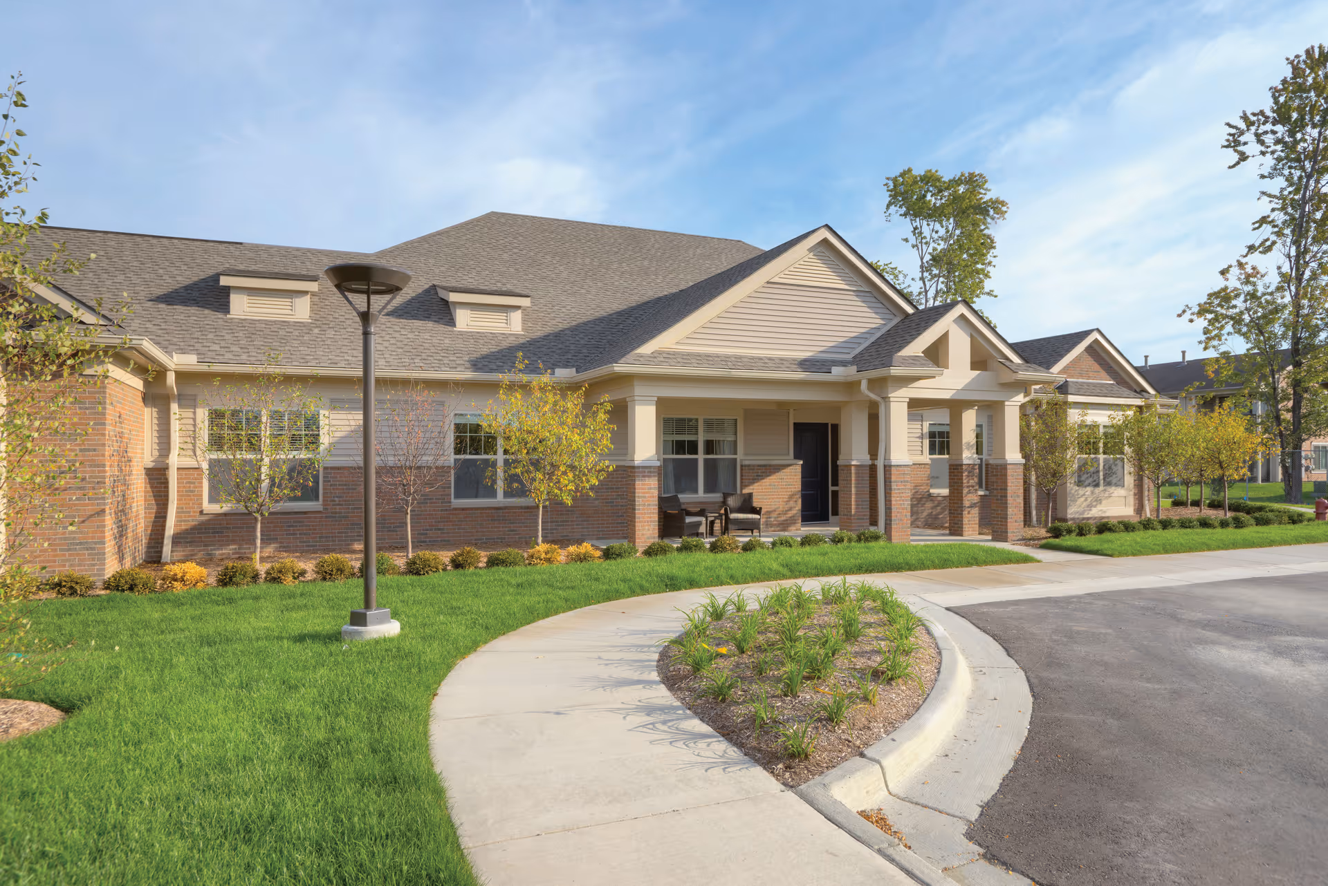 Exterior view of a single-story residential building with brick and beige siding, a gray shingled roof, and a covered entrance with columns. There is a curved concrete walkway leading to the entrance, green grass, small trees, and shrubs surrounding the building under a clear blue sky.