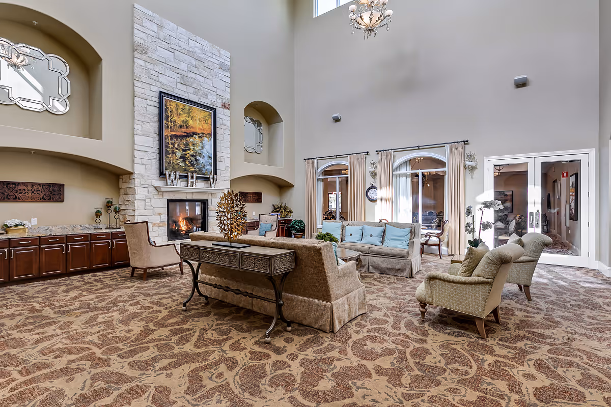 A spacious and elegant living room area with high ceilings, a stone fireplace with a painting above it, and comfortable seating including sofas and armchairs. The room features large windows with curtains, decorative mirrors, and a patterned carpet. There is a console table behind one of the sofas with a decorative sculpture on it.