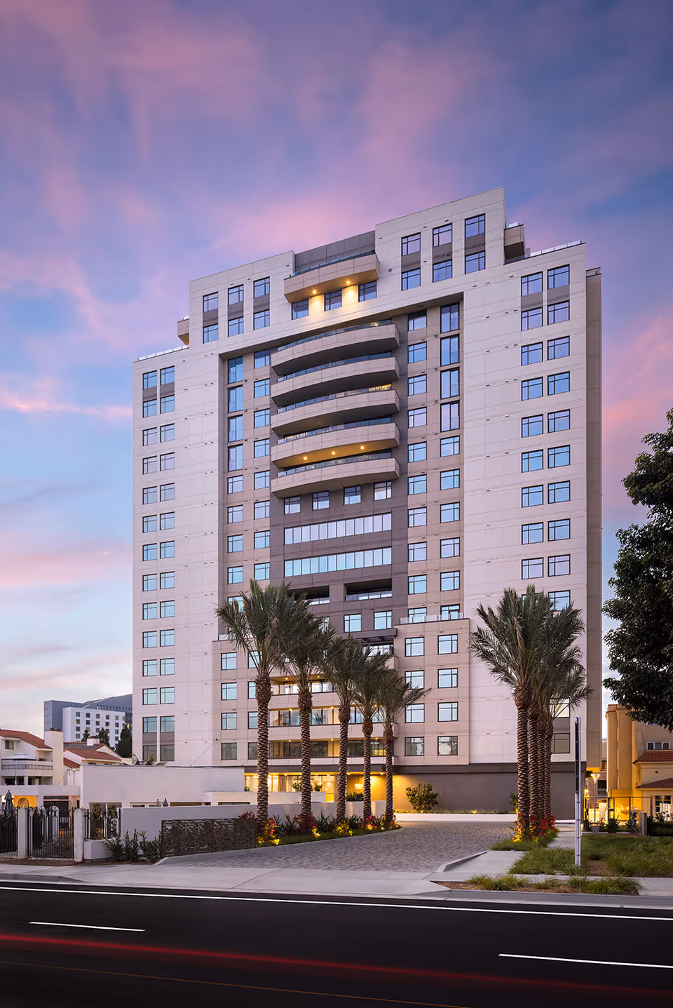 High-rise modern senior living building at dusk with balconies and palm trees lining the entrance.