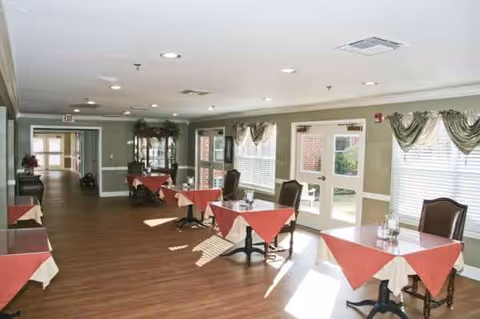 Bright dining room inside a senior living facility with red tablecloths and windows.