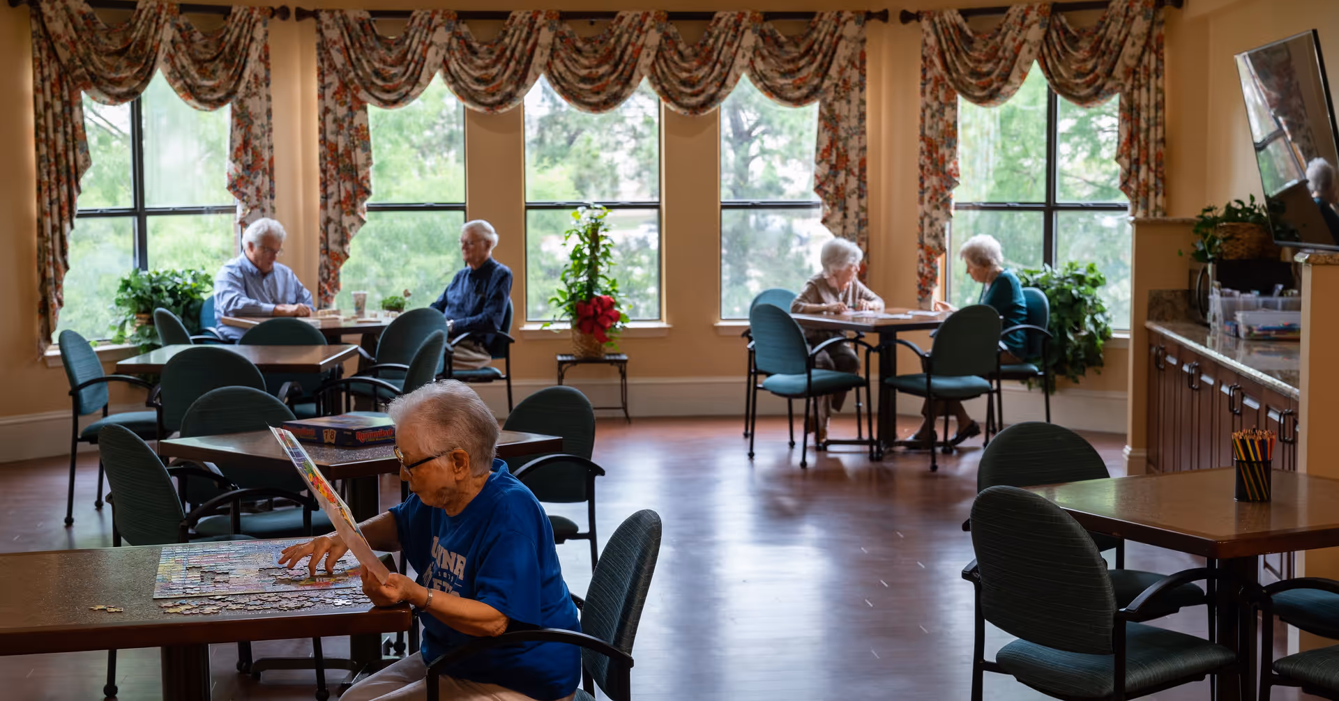 A common room in a senior living facility with several elderly people engaged in activities. One woman in the foreground is working on a jigsaw puzzle, while two pairs of seniors sit at separate tables near large windows with floral curtains, talking or playing games. The room has wooden floors, green cushioned chairs, and plants near the windows.