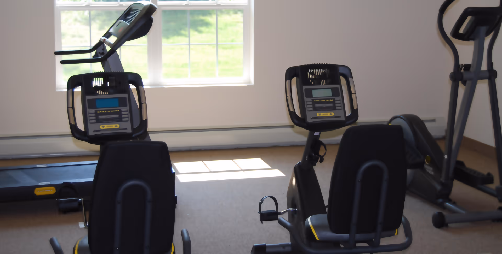 Exercise room with two recumbent stationary bikes, a treadmill, and an elliptical machine in front of a window with natural light coming in.