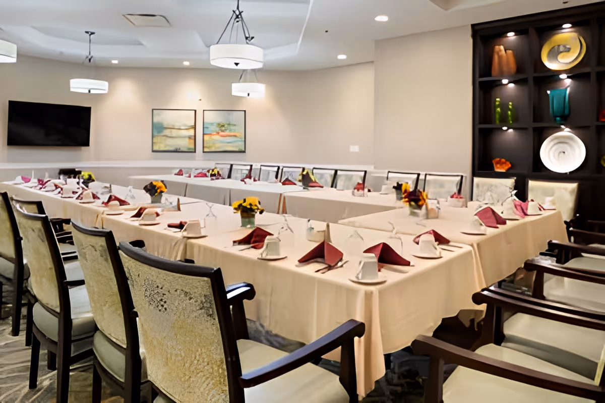 A dining room set up with tables arranged in a U-shape, covered with beige tablecloths. Each place setting includes a white cup, saucer, and a folded maroon napkin. Small flower arrangements are placed along the center of the tables. The room has cushioned chairs with wooden armrests, modern ceiling lights, a wall-mounted TV, and decorative shelves with art pieces.