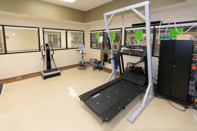 A rehabilitation gym room with exercise equipment including a treadmill with harness support, a stationary bike, and a weight scale. The room has beige walls, large windows with blinds, and a cabinet with colorful exercise balls on the right side.