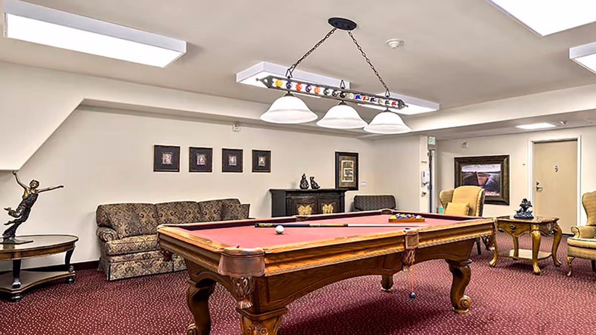A recreational room with a red felt pool table in the center, surrounded by patterned sofas and armchairs. The room has a burgundy carpet, framed pictures on the walls, a decorative statue on a side table, and a hanging light fixture above the pool table.