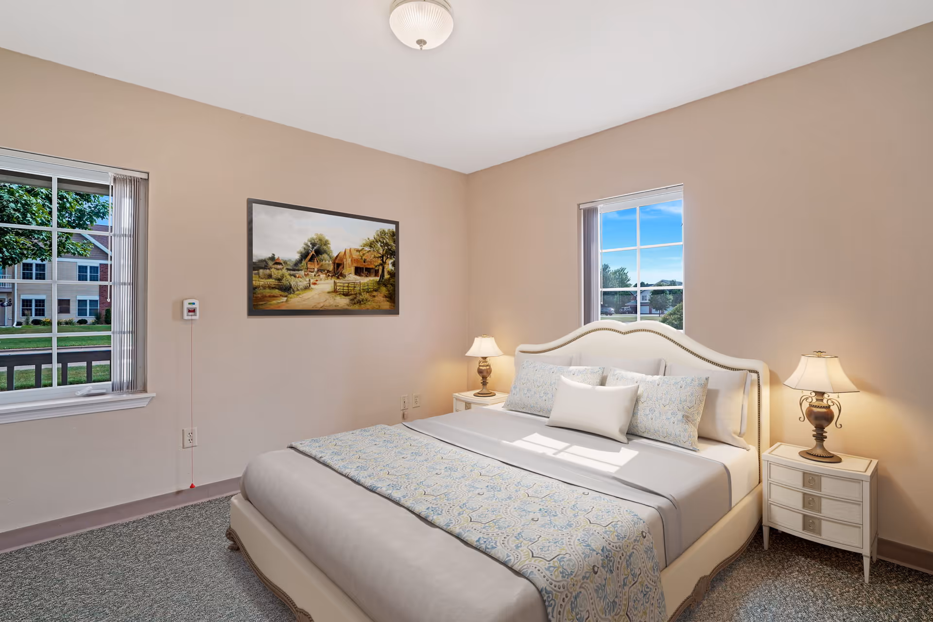 A neatly made bed with a white upholstered headboard and light blue patterned bedding in a bedroom with beige walls. There are two windows showing an outdoor view of a residential area, two bedside tables with lamps, and a framed painting of a rural scene on the wall.